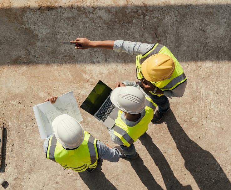 3 Männer auf einer Baustelle mit Schutzhelmen von oben Fotografiert. Der Linke hält einen Bauplan der mittlere hält einen Laptop und der rechte zeigt auf die Baustelle.