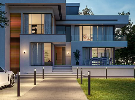 Eine moderne, mehrstöckige Villa bei Abenddämmerung, deren Einfahrt und Gartenweg von einer Reihe niedriger, schwarzer Außenleuchten beleuchtet wird, die auch als Pollerleuchten bezeichnet werden, und deren Fassade zusätzlich von zwei Wandleuchten akzentuiert wird.