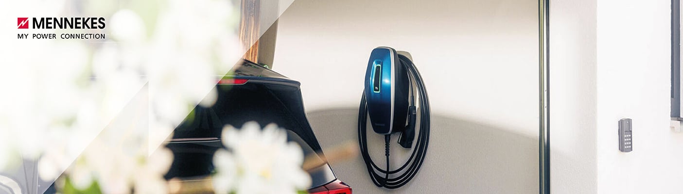  Eine moderne, blaue Ladesäule für Elektroautos hängt an der Wand eines weißen Hauses, im Vordergrund ist verschwommen ein schwarzes Auto zu sehen.