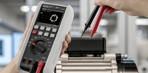 Ein Techniker in blauer Arbeitskleidung überprüft mit einer Messspitze die Elektronik eines geöffneten Gehäuses an einem Prüfplatz mit Oszilloskop.