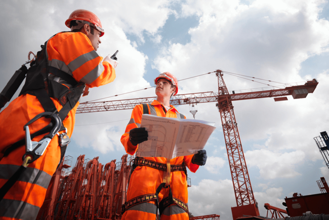 Zwei Bauarbeiter in orangefarbener Schutzkleidung auf einer Baustelle mit Kran und Bauplänen.