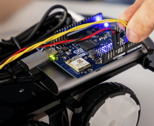 Eine Nahaufnahme zeigt eine Hand, die ein gelbes Kabel in ein blaues Arduino Uno Mikrocontroller-Board steckt, das auf einem kleinen fahrbaren Roboterchassis montiert ist.