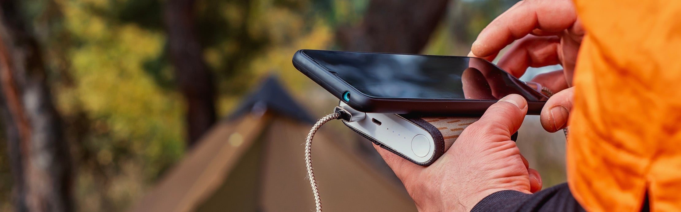 Person lädt ein Smartphone unterwegs mit einer Powerbank auf, während im Hintergrund ein Zelt im Freien steht.