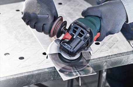 Eine Person mit grauen Schutzhandschuhen bedient einen grünen Winkelschleifer, um eine Schweißnaht an einem T-förmigen Metallteil auf einem Lochblech-Arbeitstisch zu bearbeiten, während Funken sprühen.