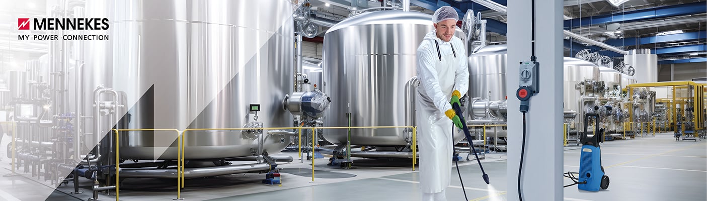  Ein Fabrikarbeiter in Schutzkleidung reinigt in einer Halle mit großen Edelstahltanks den Boden mit einem Hochdruckreiniger.