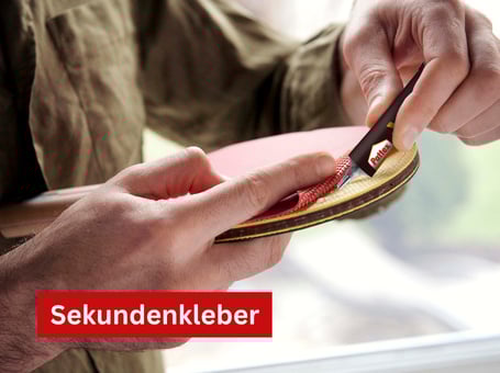 Eine Person trägt mit einer kleinen Flasche Sekundenkleber vorsichtig Klebstoff auf ein kleines, rundes Holzstück auf.
