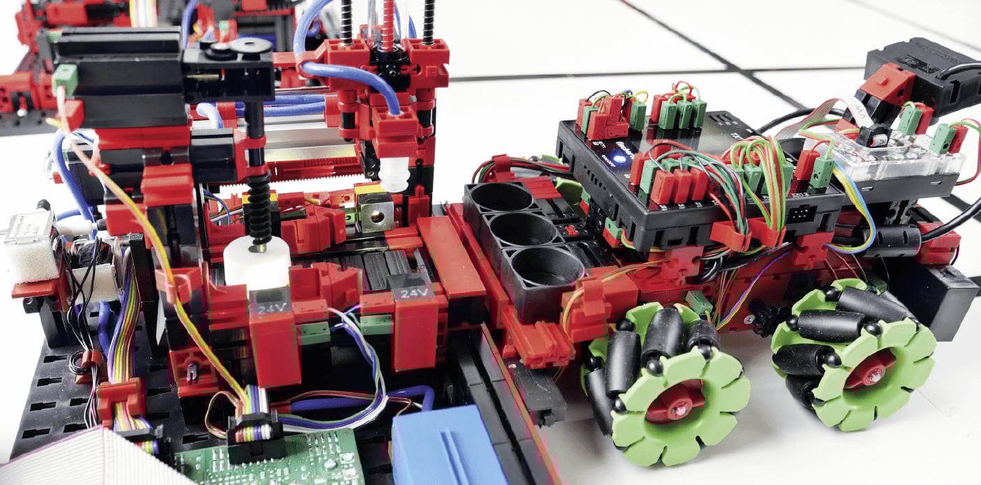 Nahaufnahme eines komplexen, modularen Technik-Aufbaus aus roten und schwarzen Elementen, an dem ein kleiner Roboter mit auffälligen grünen Mecanum-Rädern und sichtbarer Verkabelung angedockt ist.