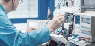 Ein Techniker in blauer Arbeitskleidung misst mit einer Prüfspitze an einer komplexen elektronischen Schaltung, die von verschiedenen Messgeräten wie Oszilloskopen und Netzteilen umgeben ist.