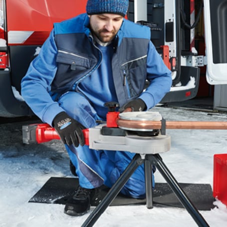 Ein Handwerker kniet im Schnee vor einem Lieferwagen und biegt ein Kupferrohr mit einer akkubetriebenen Rohrbiegemaschine auf einem Stativ.