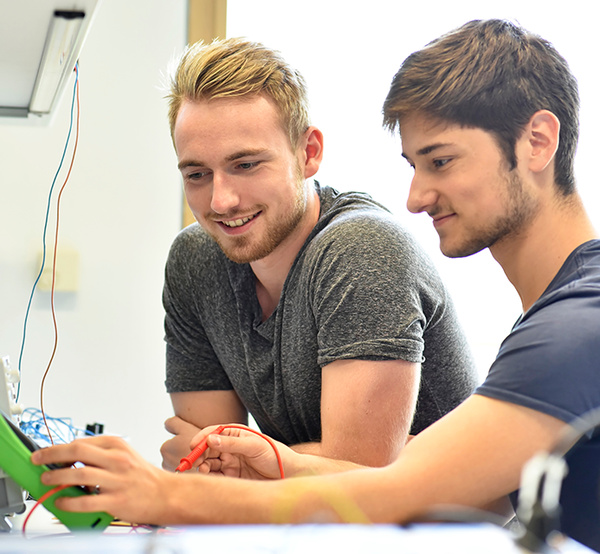 Zwei junge Männer prüfen elektrische Schaltungen mit einem Messgerät im Labor oder Workshop.