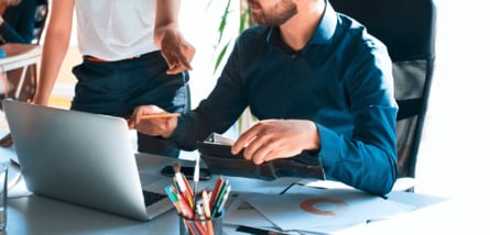 Ein Mann im blauen Hemd arbeitet an einem Laptop und hält einen Ordner, während eine weitere Person neben ihm steht und auf den Bildschirm deutet.