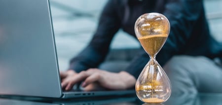 Eine Sanduhr mit goldfarbenem Sand steht auf einem Tisch vor einer Person, die an einem Laptop arbeitet.