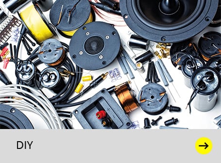 Das Bild zeigt eine Vielzahl von elektronischen Bauteilen für den Lautsprecherbau, darunter Spulen, Kondensatoren, Kabel, Lautsprecherchassis und Hochtöner, unordentlich auf einer hellen Oberfläche verteilt, unterhalb dessen sich ein grauer Balken mit der schwarzen Aufschrift "DIY" und einem gelben Pfeil nach rechts befindet.