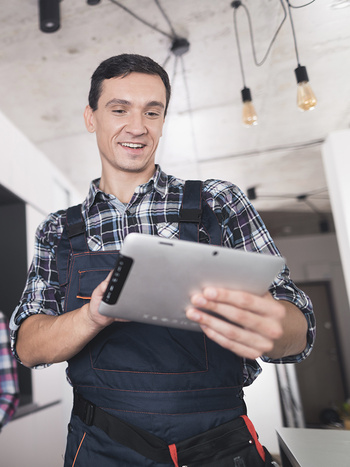 Ein lächelnder Handwerker in Arbeitskleidung betrachtet ein Tablet, während er an einer Installation arbeitet.