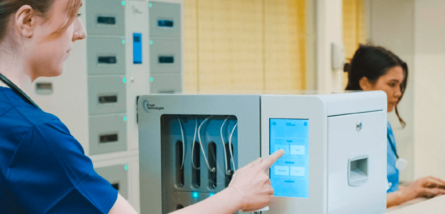 Eine Pflegekraft in blauer Dienstkleidung bedient den Touchscreen einer modernen, weißen Ladestation für medizinische Geräte von Power Technologies, während im Hintergrund eine weitere Person am Computer arbeitet.