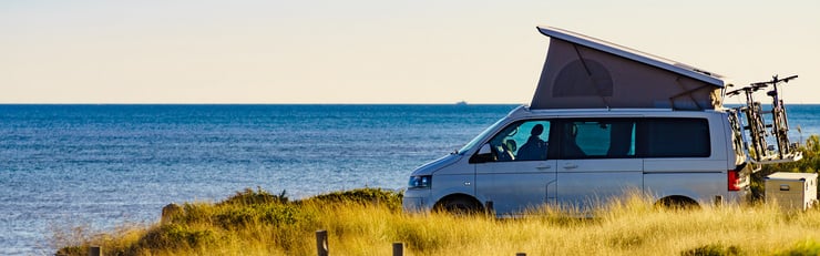 Ein Campingbus mit ausgeklapptem Dach steht am Meer, im Hintergrund ist der Horizont sichtbar.
