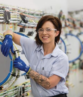 Das Bild zeigt eine Arbeiterin mit Ansell Arbeitshandschuhen, die an eine Maschine angelehnt in einer Industriehalle steht.