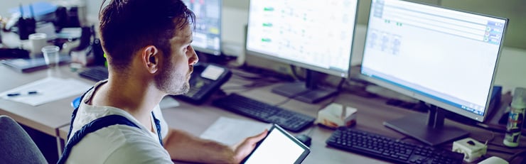 Ein Techniker in Arbeitskleidung sitzt in einem Kontrollraum vor mehreren Monitoren mit komplexen Systemanzeigen. In der einen Hand hält er ein Tablet, mit der anderen bedient er eine Computermaus.