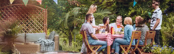 Gruppe von Freunden sitzt an einem gedeckten Tisch im Garten, trinkt und lacht bei einem sommerlichen Beisammensein unter bunten Wimpeln.
