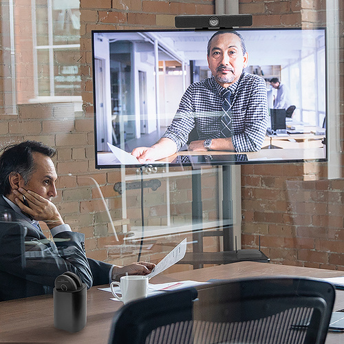 Ein Mann nimmt an einer Videokonferenz in einem modernen Büro teil, während ein anderer Gesprächspartner auf einem Bildschirm zu sehen ist.