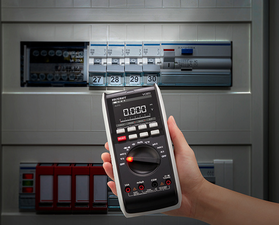 Hand hält Voltcraft VC851 Multimeter vor einem Sicherungskasten und misst Spannung.
