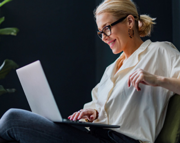 Eine lächelnde Frau mit Brille sitzt entspannt und arbeitet an einem Laptop.