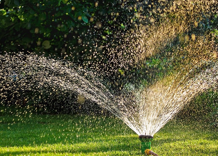 Rasensprenger bewässert bei Sonnenschein einen grünen Gartenrasen mit weit auffächerndem Wasserstrahl.