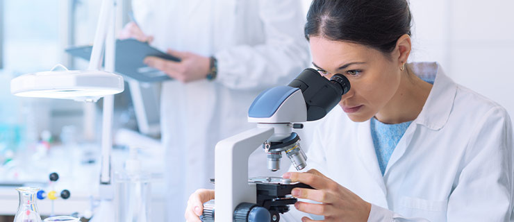 Wissenschaftlerin im weissen Laborkittel blickt durch ein Mikroskop, während ein Kollege im Hintergrund Notizen auf einem Klemmbrett macht