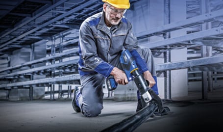 Ein Arbeiter in Schutzkleidung und Helm kniet auf einem Betonboden und schneidet ein dickes Industriekabel mit einem hydraulischen Akkuschneider.