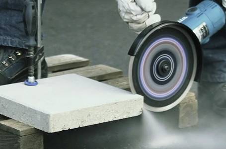 Eine Person schneidet eine eingespannte Betonplatte mit einem blauen Winkelschleifer, der mit einer Diamanttrennscheibe ausgestattet ist, wobei feiner Staub aufgewirbelt wird.