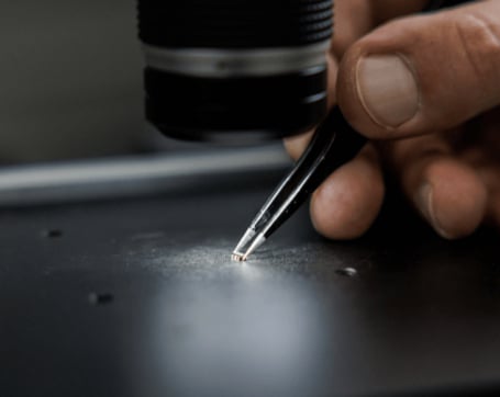 Nahaufnahme einer Hand, die mit einer Pinzette ein winziges elektronisches Bauteil unter einem Mikroskop auf einer dunklen Arbeitsfläche präzise platziert.