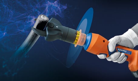 Eine behandschuhte Hand hält ein orange-blaues, isoliertes hydraulisches Schneidwerkzeug, das gerade ein kunststoffummanteltes Kabel durchtrennt, mit abstrakten Lichteffekten im Hintergrund.