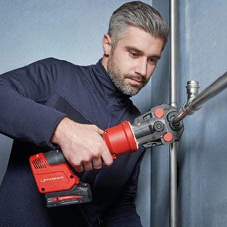 Ein Handwerker verbindet zwei Metallrohre an einem T-Stück mithilfe einer roten, akkubetriebenen Rohrpressmaschine.
