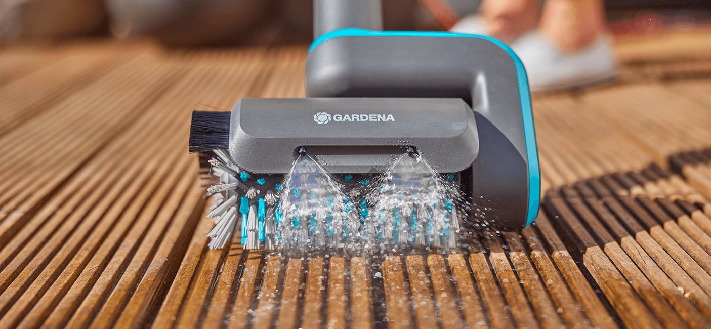Ein Gardena Reinigungsbürste mit Wasserstrahlen säubert eine Holzterrasse effektiv.