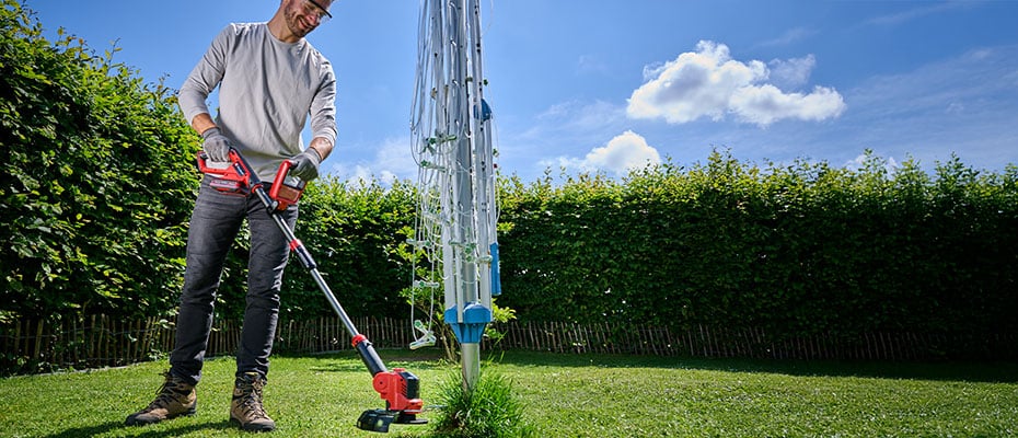  Ein Gärtner in Latzhose und Hemd hält in einem grünen Garten einen roten Einhell Rasenmäher, während er Rasen mäht und lächelt.