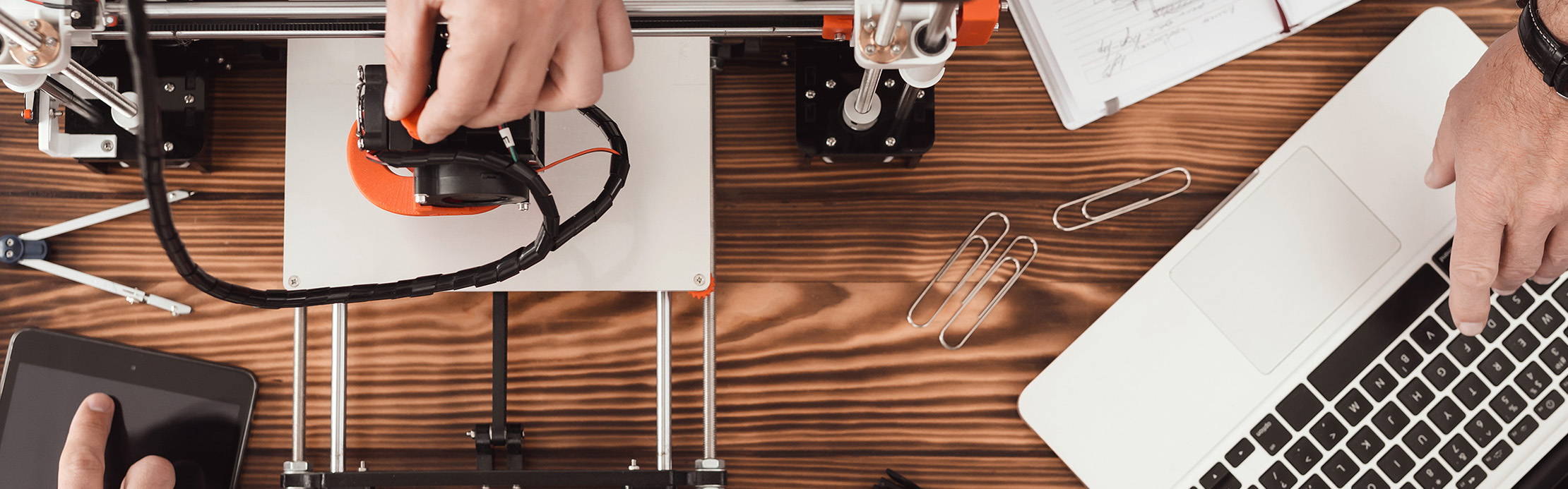 Draufsicht auf einen Arbeitsplatz mit 3D-Drucker, Laptop, technischen Skizzen und Arbeitsmaterialien für den Prototypenbau.