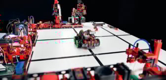 Ein kleiner, aus roten und schwarzen Bauteilen konstruierter Roboter fährt auf einer weißen Fläche mit schwarzen Linien zwischen verschiedenen technischen Fischertechnik-Modellen umher.