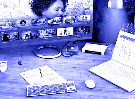 Eine monochrome, blau eingefärbte Ansicht eines Schreibtisches mit einer Tastatur und Maus im Vordergrund, einem Laptop auf der rechten Seite und einem großen Monitor im Hintergrund, der ein Streaming-Menü anzeigt und über kleine KVM-Umschaltboxen mit den Geräten verbunden ist.
