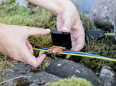 Zwei Hände platzieren eine elektrische Kabelverbindung mit Verbindungsklemmen in eine schwarze, mit Gel gefüllte Schutzbox, die im Außenbereich auf moosigem Untergrund liegt.
