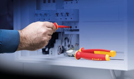 Nahaufnahme einer Hand, die mit einem rot-gelb isolierten VDE-Schraubendreher an einem elektrischen Bauteil arbeitet, mit einer passenden VDE-Kombizange im Vordergrund.
