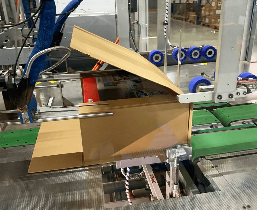  Eine braune Pappverpackung wird von einem Förderband in einer industriellen Produktionsanlage zusammengefaltet.