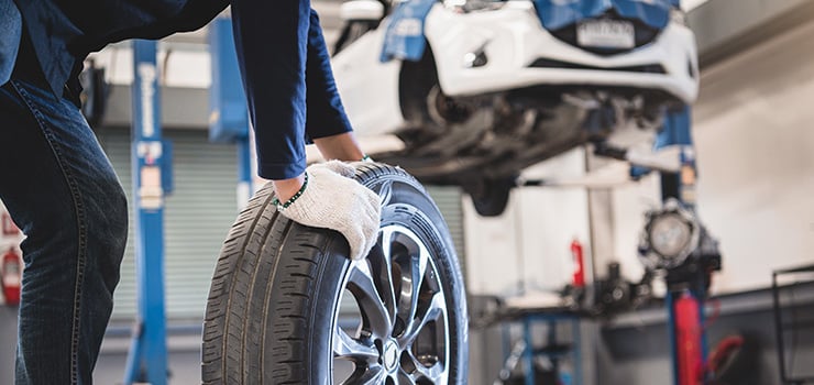 Eine Person mit Arbeitshandschuhen hebt einen Autoreifen in einer Werkstatt an, während ein Fahrzeug auf einer Hebebühne steht.