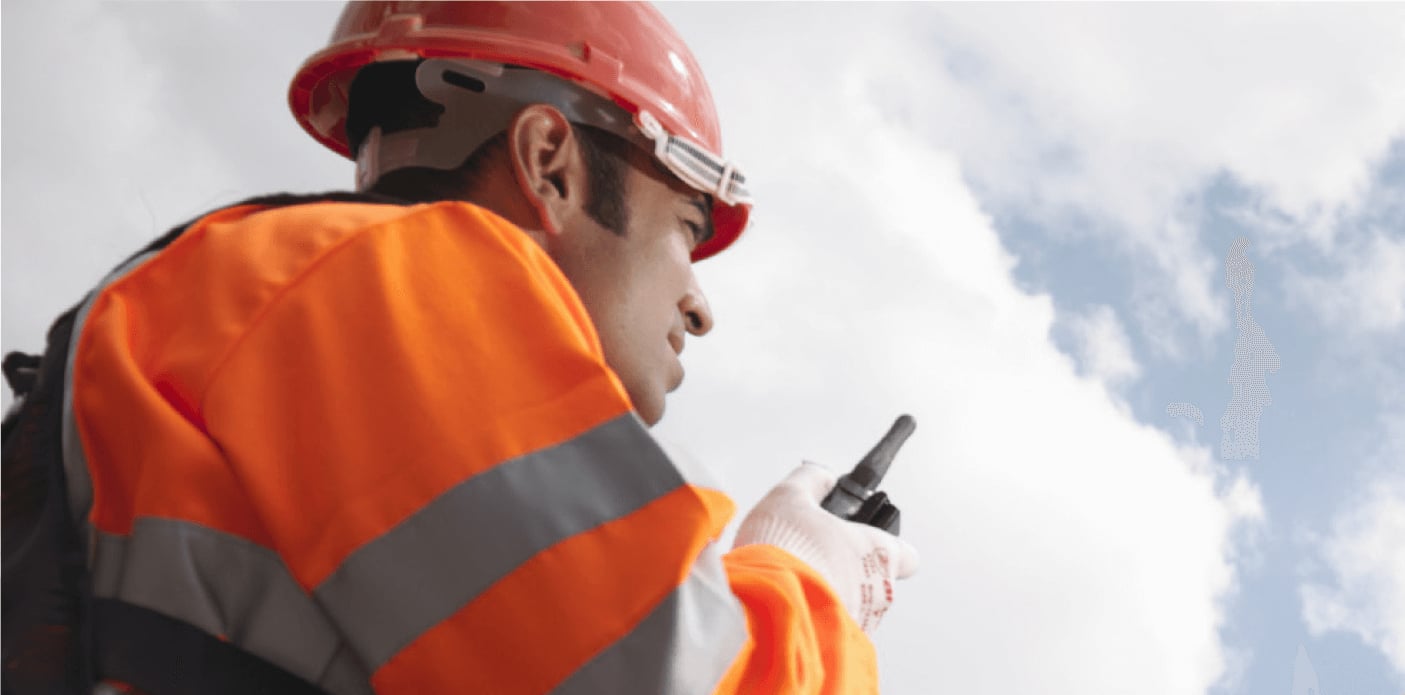 Ein Arbeiter in orangefarbener Warnkleidung und rotem Schutzhelm spricht vor einem bewölkten Himmel in ein Funkgerät.