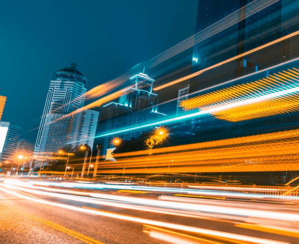 Nächtliche Langzeitbelichtung einer städtischen Hauptstraße mit dynamischen, leuchtend orangefarbenen und blauen Lichtspuren vorbeifahrender Fahrzeuge vor einer Kulisse aus beleuchteten Hochhäusern.