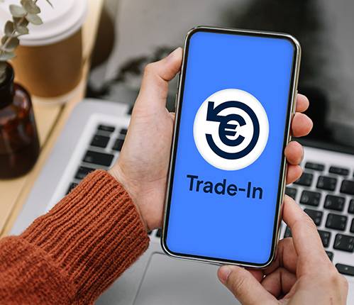 Gerne, hier eine kürzere Version:  Hände halten ein Smartphone mit einem "Trade-In"-Logo (Recycle-Symbol mit Euro-Zeichen) auf blauem Hintergrund. Im unscharfen Hintergrund sind ein Laptop und eine Tasse zu sehen.