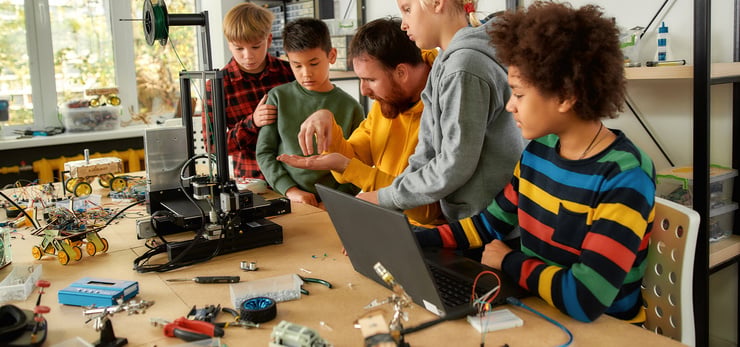 Lehrer erklärt einer Gruppe von vier Kindern an einem Tisch mit 3D-Drucker und Laptop elektronische Bauteile.