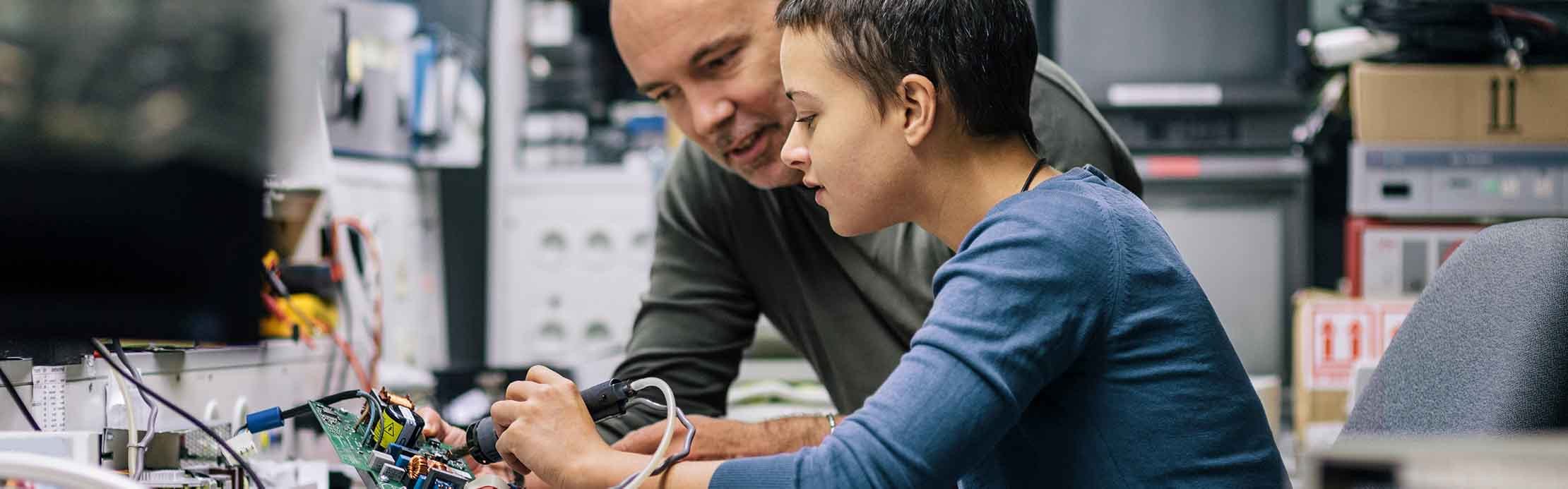 Neben einem Mann sitzt eine Frau, die einen Lötkolben und ein Motherboard in die Hände hält und lötet.