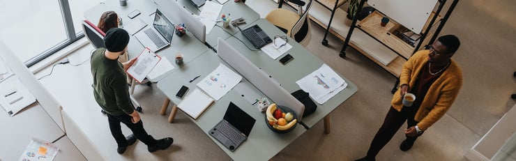 Zwei Personen arbeiten gemeinsam an einem Schreibtisch im Büro und besprechen Inhalte auf einem Laptop, während im Hintergrund eine weitere Person an einem Computer sitzt.