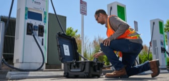 Ein Techniker in Warnweste kniet an einer E-Auto-Ladestation neben einem offenen Werkzeugkoffer, um Wartungsarbeiten oder Tests an der Ladesäule durchzuführen.