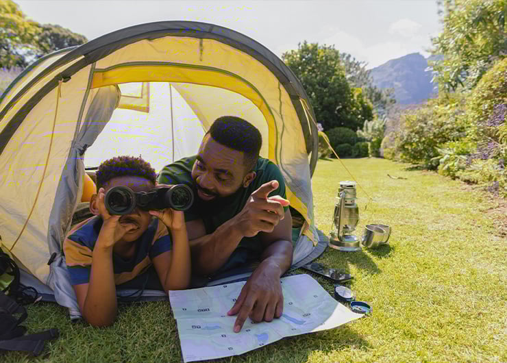 Ein Vater und sein Sohn liegen vor einem Zelt auf einer Wiese, schauen durch ein Fernglas und betrachten gemeinsam eine Karte.